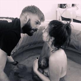A mother is sat in a birth pool holding her newborn baby and reaches up to kiss her partner