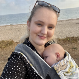 Independent Midwife Mollie may standing on the beach with a baby in a sling 